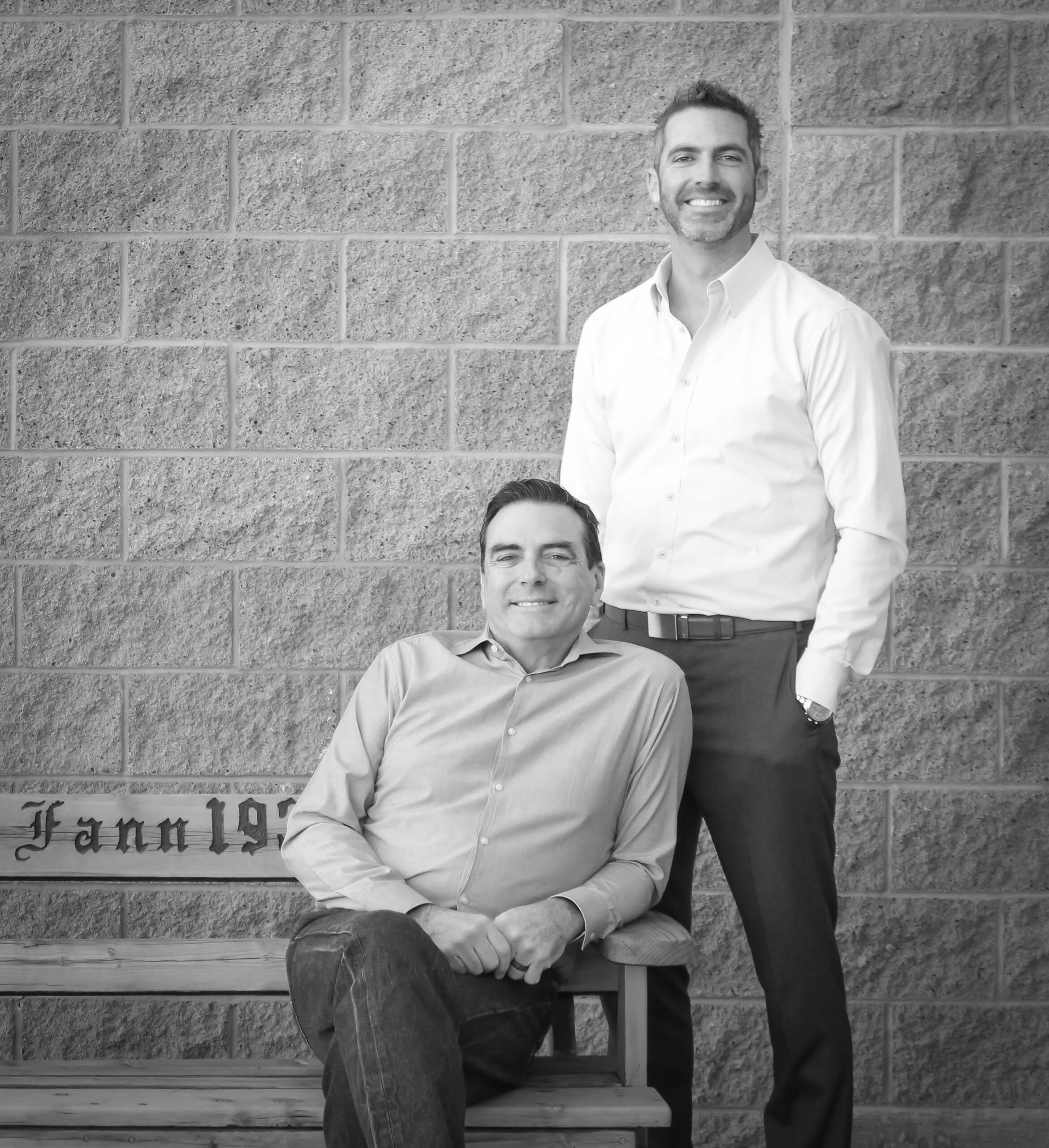 Two men sitting on a bench in front of a brick wall, showcasing Fann Contracting.