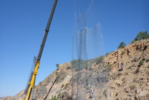 A crane lifts a large net over the side of a mountain.